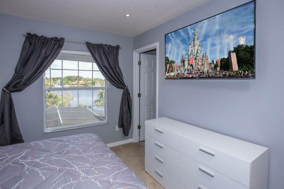Bright and airy, this bedroom features soft gray walls complemented by a cozy bedspread and sleek white dresser. Enjoy the scenic view from the large window, framed by elegant dark curtains, while a modern TV adds entertainment.
