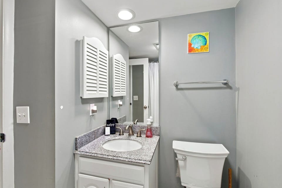 Speckled stone counters and crisp white cabinetry create a clean, refreshing atmosphere for your morning routine. A tall mirror reflects the calming gray walls and vibrant artwork, making this tidy bathroom feel airy and bright.
