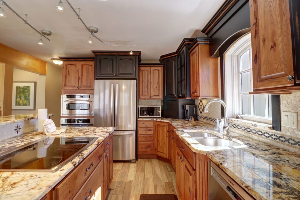 A modern kitchen featuring a stainless steel refrigerator, granite countertops, wooden cabinets, an electric stovetop, a dishwasher, and a window above the sink. Track lighting illuminates the space.