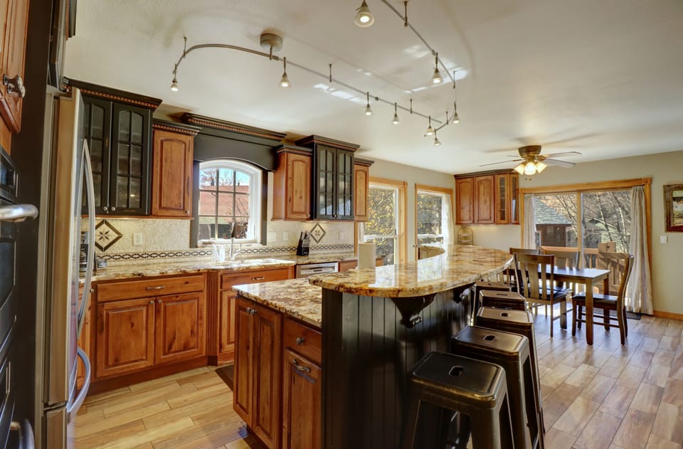 A modern kitchen with wooden cabinets, marble countertops, and bar seating. The space includes track lighting, a ceiling fan, and a dining area with a table and chairs near large windows.