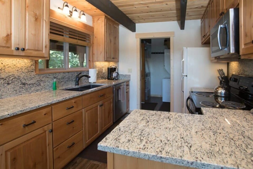The spacious and open kitchen is fully equipped with basic cookware and features stainless steel appliances, spacious granite counters, and an over-the-sink window with views over the surrounding forest.