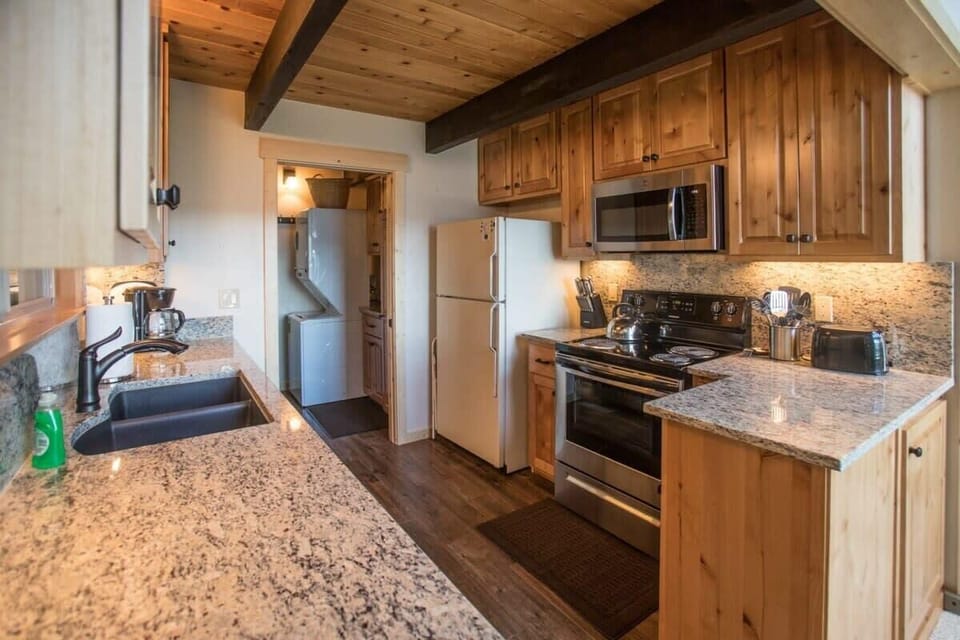 The spacious and open kitchen is fully equipped with basic cookware and features stainless steel appliances, spacious granite counters, and an over-the-sink window with views over the surrounding forest.