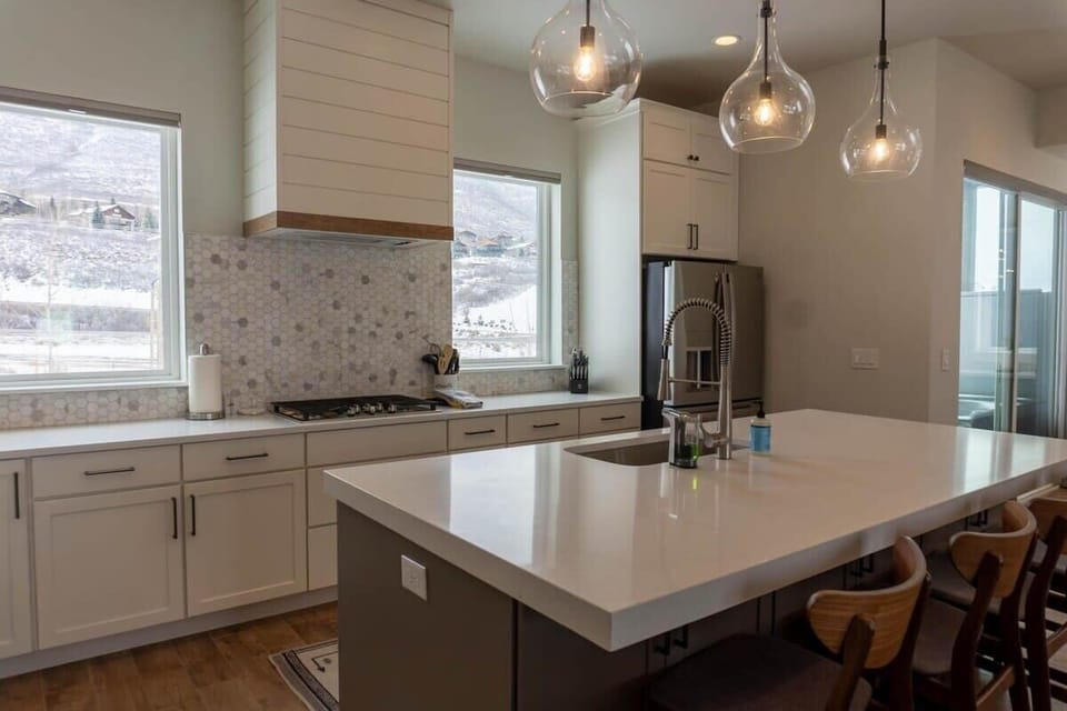 The kitchen is a bright and beautiful space featuring white cabinetry, white countertops, a tiled backsplash, stainless steel appliances, and a large kitchen bar with four barstools.