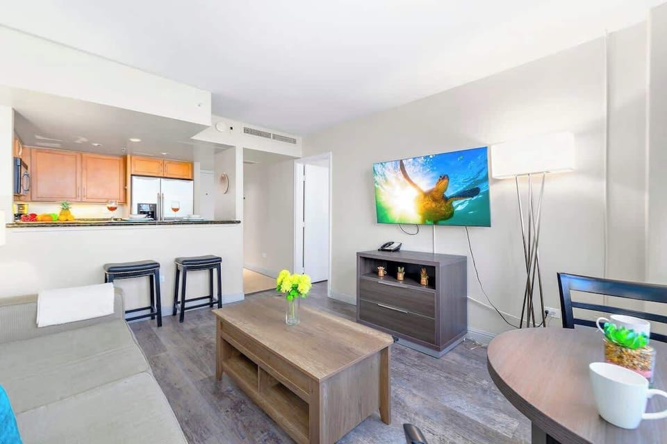 Living room with dining table for up to two people and countertop with 2 bar stools, and a flatscreen TV. 