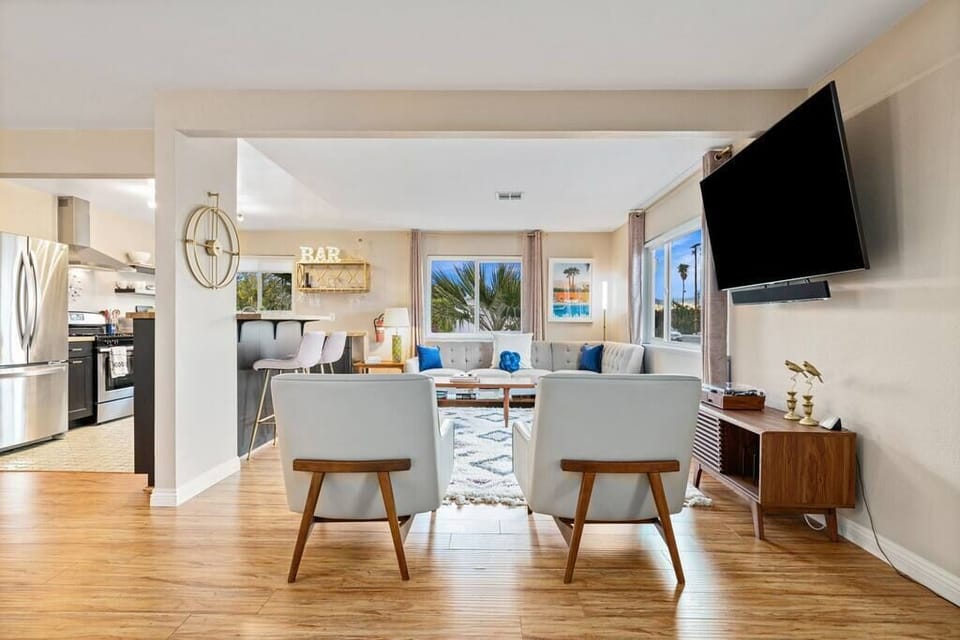 Living room with mid-century lounge chairs, wall-mounted TV, bar nook, and palm-framed windows — the group hangout hub