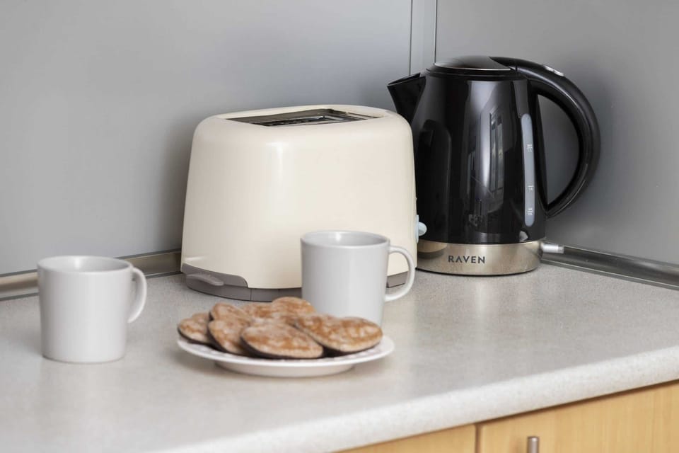A countertop with a kettle, toaster, mugs, and snacks.
