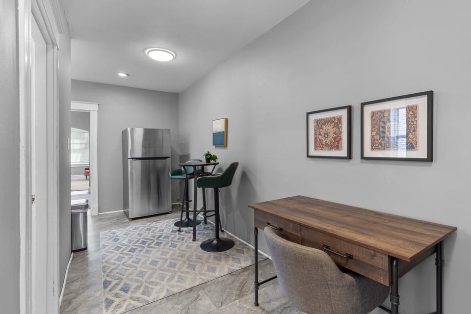 A dedicated wood desk with upholstered office chair shares the tiled hall with a stainless refrigerator, round high table, two green stools, patterned runner, and doorway toward the living room.