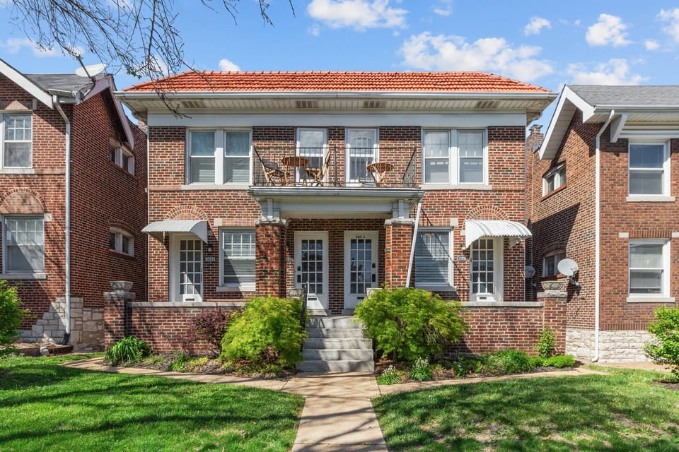 The daytime front exterior shows a two-story brick residential building with central entry steps, covered porch, upper balcony, striped door awnings, landscaped beds, lawn, and neighboring homes.