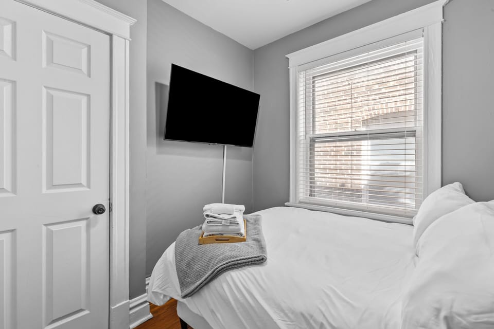 This reverse second-bedroom view shows the white-dressed bed, wall-mounted TV, large window, closet door, gray knit throw, folded towels, and hardwood floors.