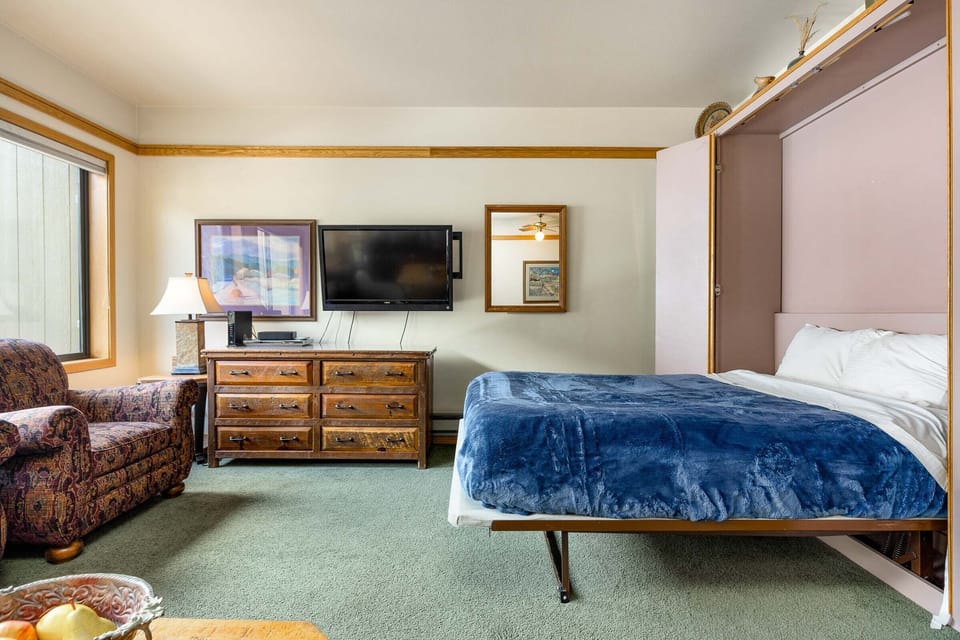 A hotel room with a queen-sized bed, wooden dresser with a flat-screen TV, a patterned armchair, and a window. Fruit and a lamp are on the table in the foreground.