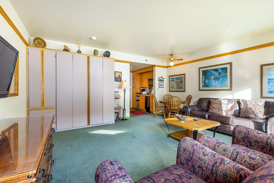 Spacious living room with green carpet, two armchairs, a sofa, coffee table, and a wall-mounted TV. A partial view of the kitchen and dining area with a table and chairs can be seen in the background.
