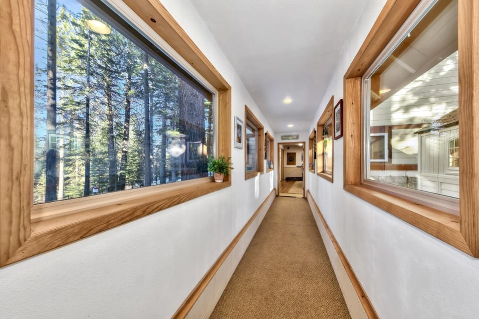 Long narrow hallway with beige carpet, white walls, natural wood window frames, and views of forest outside.