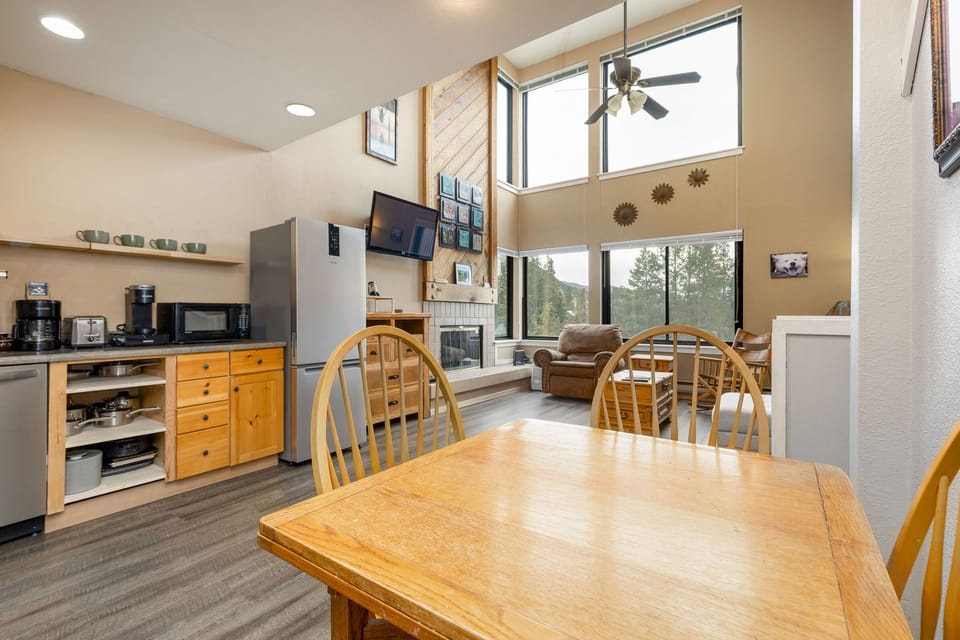 Open-concept kitchen and living room with wooden furniture, high ceilings, large windows, ceiling fan, refrigerator, stove, coffee maker, and a television mounted above a fireplace.