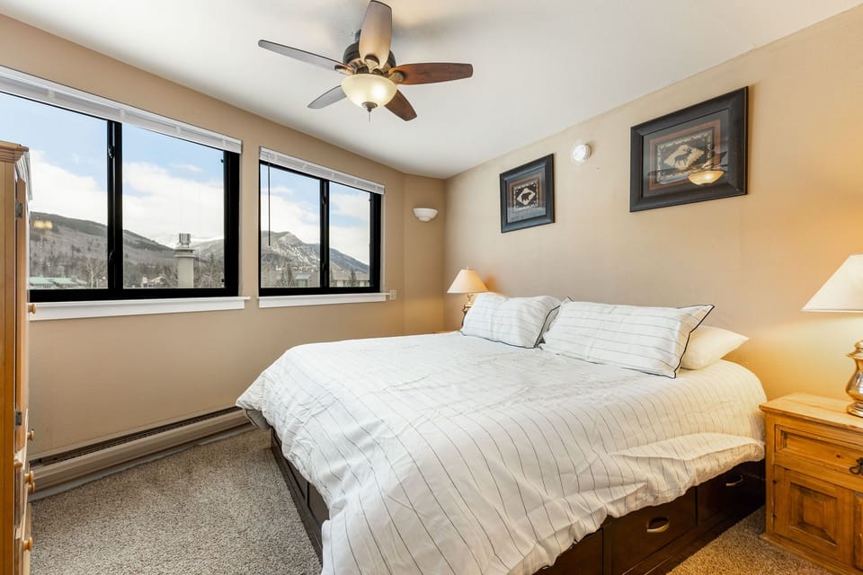 Bedroom with a large bed, white-striped bedding, two nightstands with lamps, framed pictures on beige walls, three windows with mountain views, and a ceiling fan.