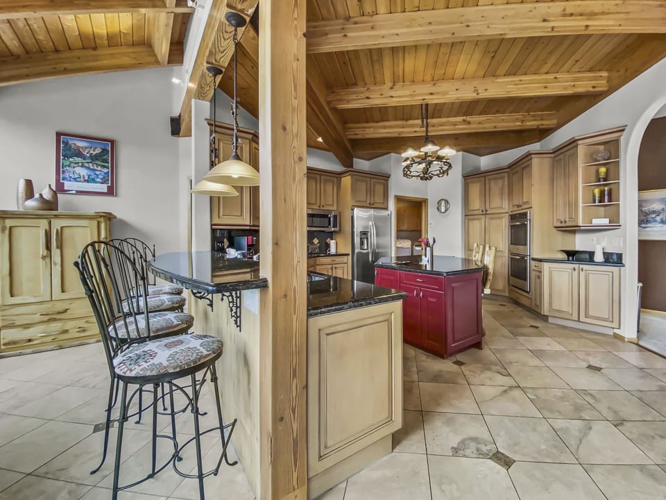 A spacious kitchen with wooden cabinetry, a central island, and a breakfast bar with high chairs. The ceiling features exposed wooden beams, and the floor is tiled. A refrigerator and oven are visible.