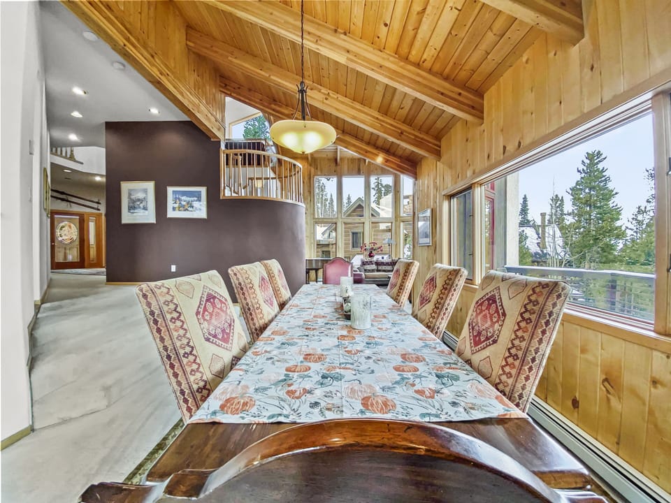 A dining area with a wooden ceiling and walls. The table is set with a patterned runner and six upholstered chairs, overlooking large windows with a view of trees.
