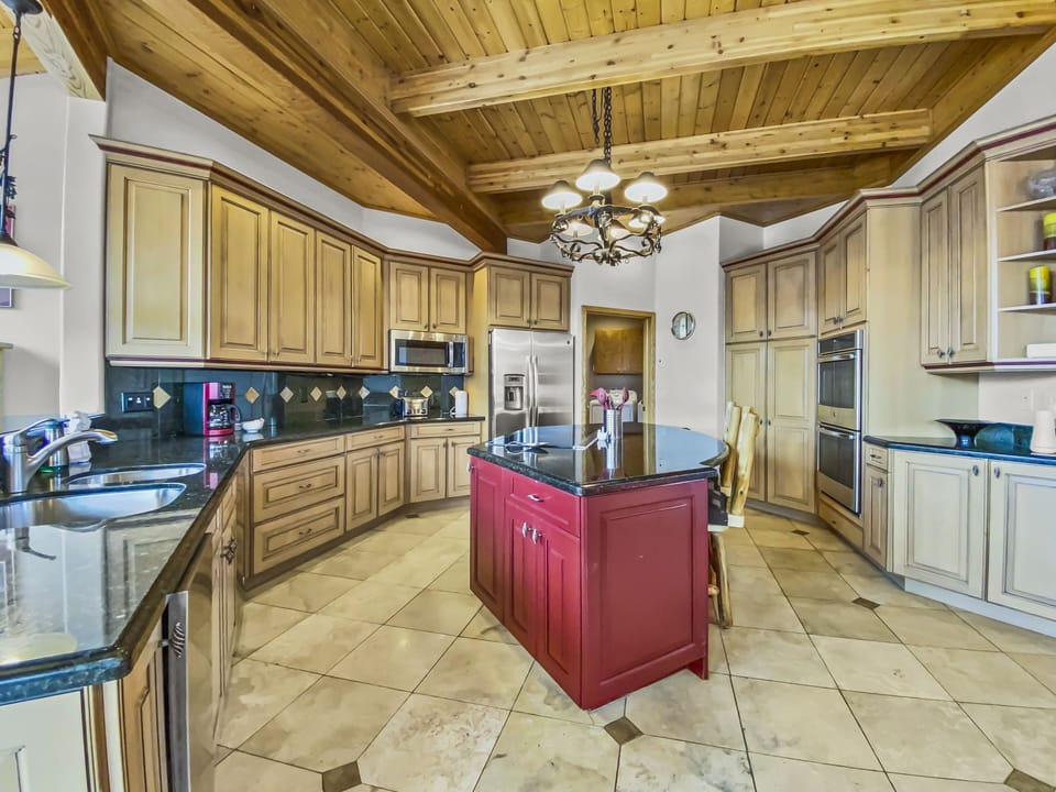 A spacious kitchen with wooden cabinets, a red island, stainless steel appliances, tiled floor, and a wooden beamed ceiling. There is a light fixture hanging above the island.
