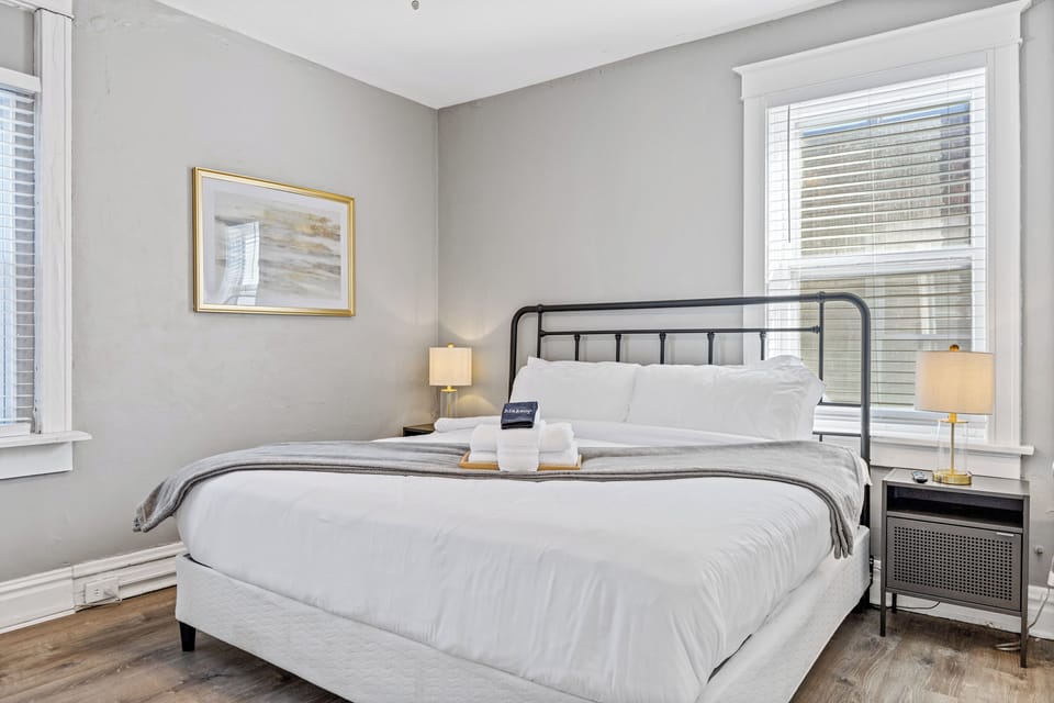 The primary bedroom features a large white-dressed bed, black metal headboard, gray throw, folded towels, matching nightstands and lamps, two windows, framed artwork, and gray flooring.