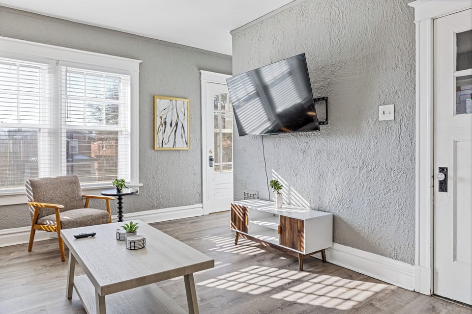 This living-room angle shows the wall-mounted TV, upholstered accent chair, white coffee table, wood-and-white media console, large windows, glass-paneled door, and gray flooring.