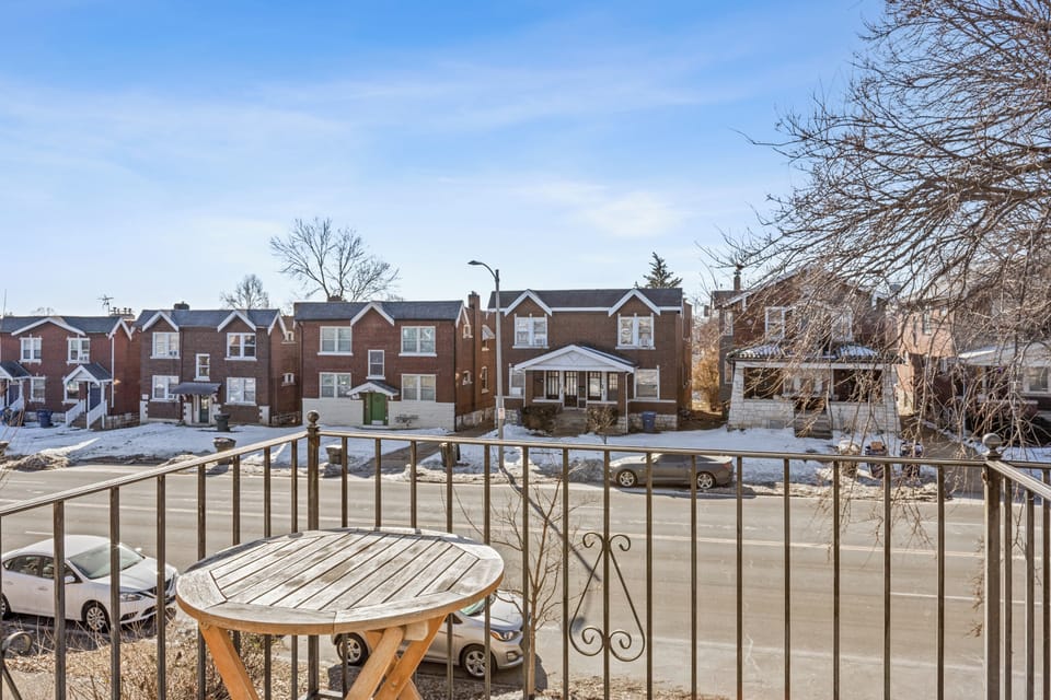 The upper balcony includes a round wood table and black metal railing with a daytime winter view across the residential street toward neighboring brick homes.