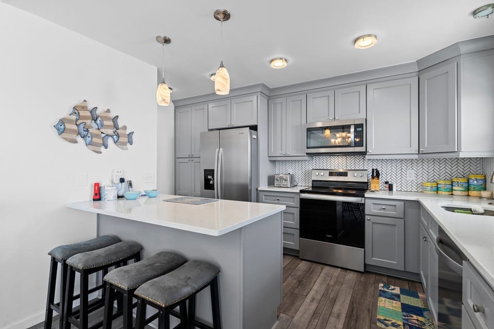 Gray coastal kitchen with island seating, stainless appliances, white counters, herringbone backsplash, pendant lights, fish wall art, and beachy decor.