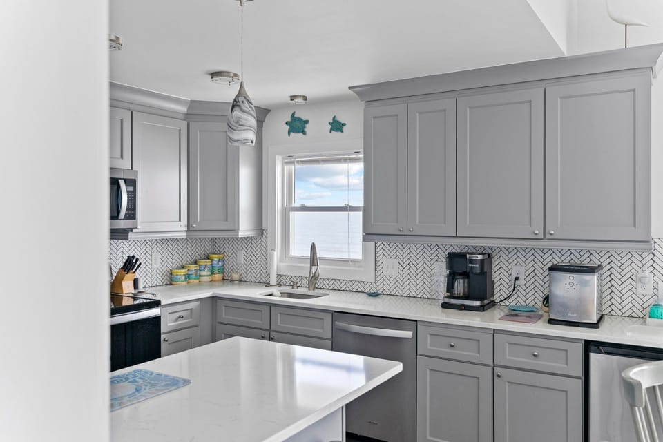 Gray coastal kitchen with island seating, stainless appliances, white counters, herringbone backsplash, pendant lights, fish wall art, and beachy decor.
