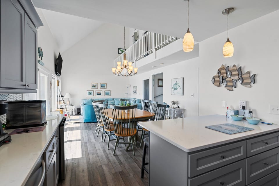 Gray coastal kitchen with island seating, stainless appliances, white counters, herringbone backsplash, pendant lights, fish wall art, and beachy decor.