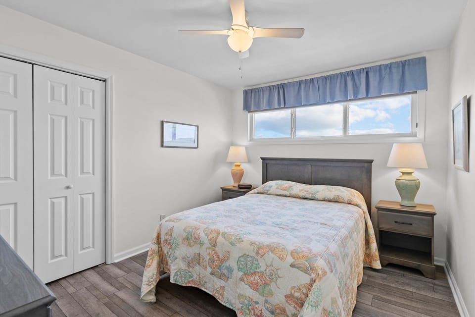 Oak Island bedroom with two twin beds, seashell bedding, dark headboards, ceiling fan, wide window, closet doors, framed art, lamp, books, and wood floors.