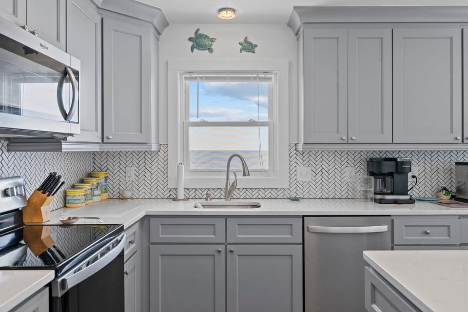 Gray coastal kitchen with island seating, stainless appliances, white counters, herringbone backsplash, pendant lights, fish wall art, and beachy decor.