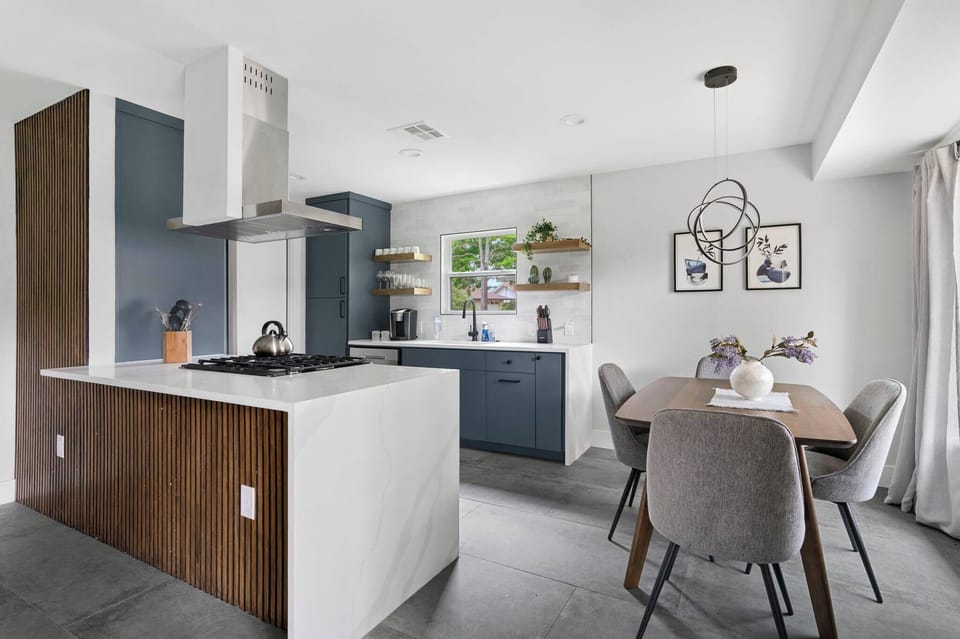 Modern kitchen with sleek island, stainless appliances, and open shelving — flowing seamlessly into a bright dining space with stylish seating