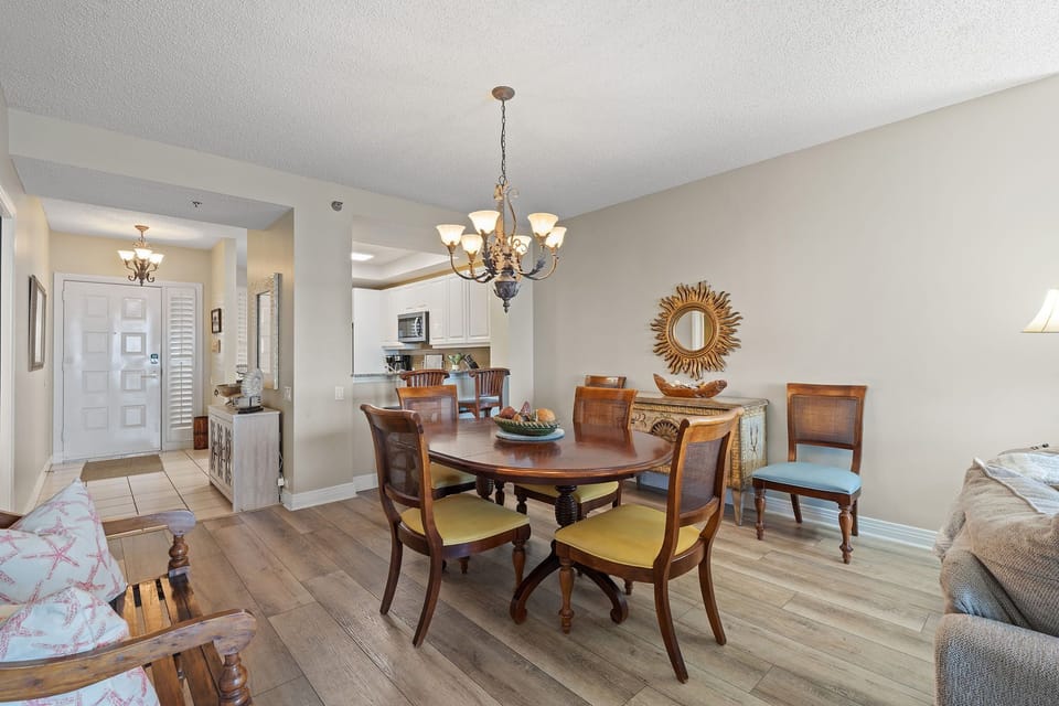 Beach Colony T15B- Step into your inviting dining area with warm wood floors and elegant furnishings. Gather around the oval table under soft chandelier light, with your open kitchen just steps away for easy entertaining.