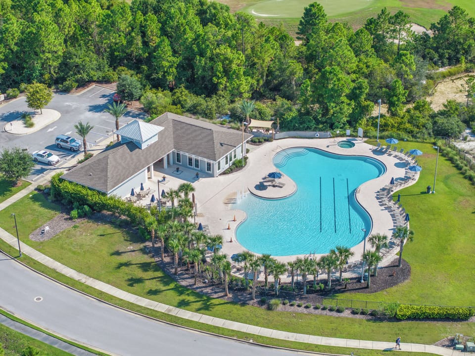 Lost Key Community Aerial view showcasing the large freeform swimming pool with adjacent clubhouse, surrounded by lush landscaping and bordered by a golf course. The community amenity area includes ample parking and is nestled among towering trees.