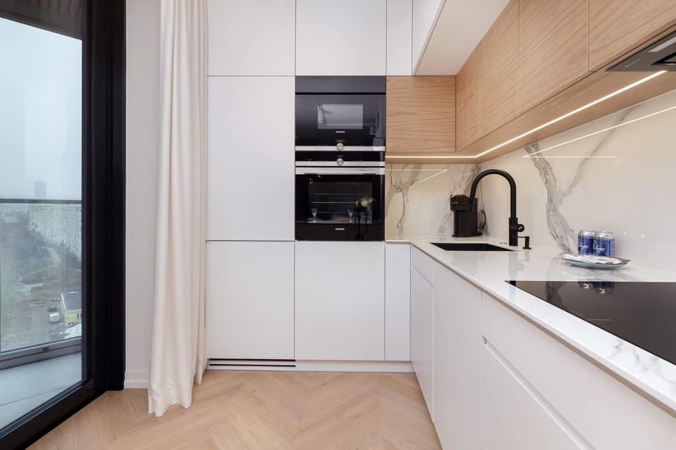 A modern kitchen with white cabinets and wooden accents, including built-in appliances and a glass door leading to the balcony.
