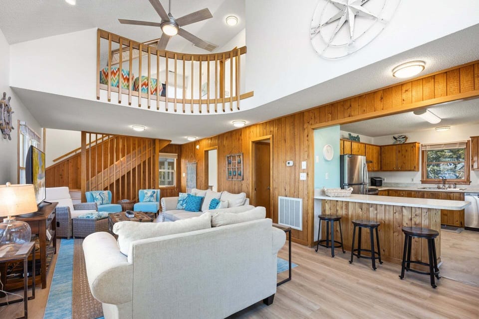 The spacious living area features high ceilings, bright natural light, and coastal-inspired décor, creating a warm and inviting place to relax around the flat-screen TV.