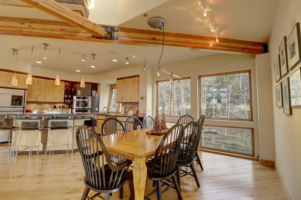 A modern kitchen and dining area with a wooden table and black chairs. The kitchen features wooden cabinets, stainless steel appliances, and pendant lighting. Large windows provide natural light.