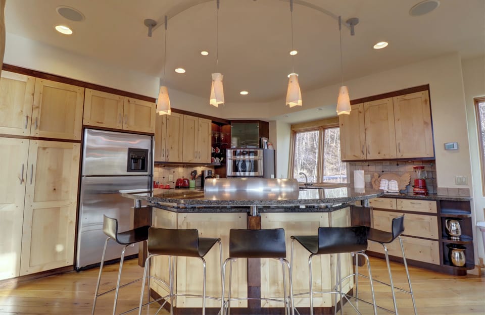 Modern kitchen featuring wooden cabinetry, stainless steel appliances, a central island with black barstools, and pendant lighting above.
