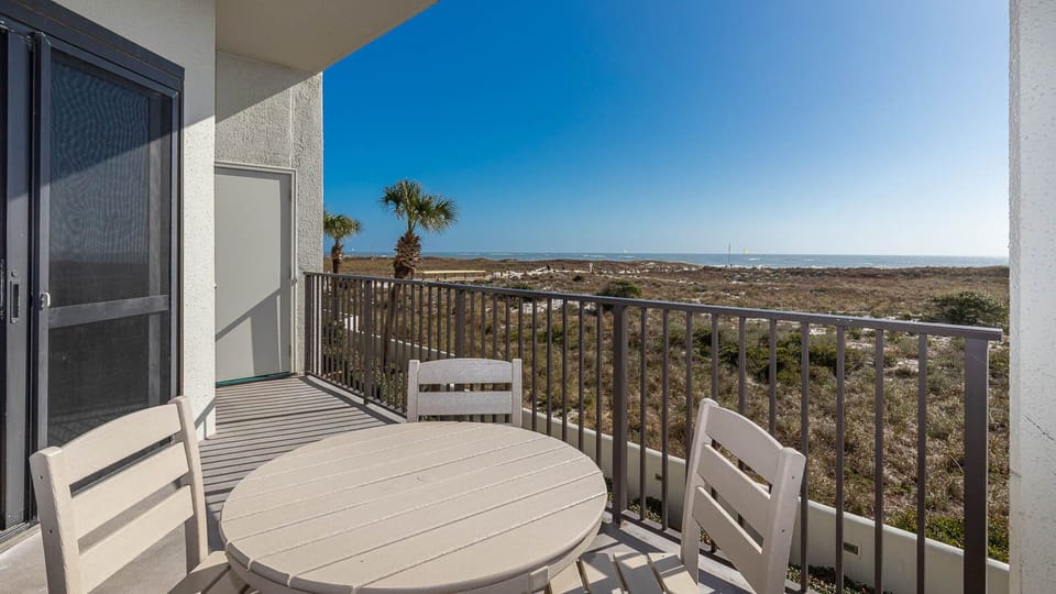 Balcony with a round table and chairs overlooks lush greenery and the tranquil gulf, perfect for enjoying the view