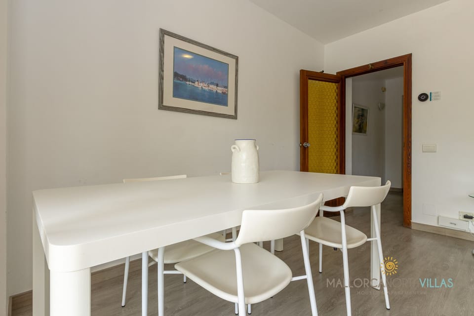 Indoor dining area with white table and chairs, decorative jug, nautical artwork and yellow glass door to the hallway. Laminate flooring.