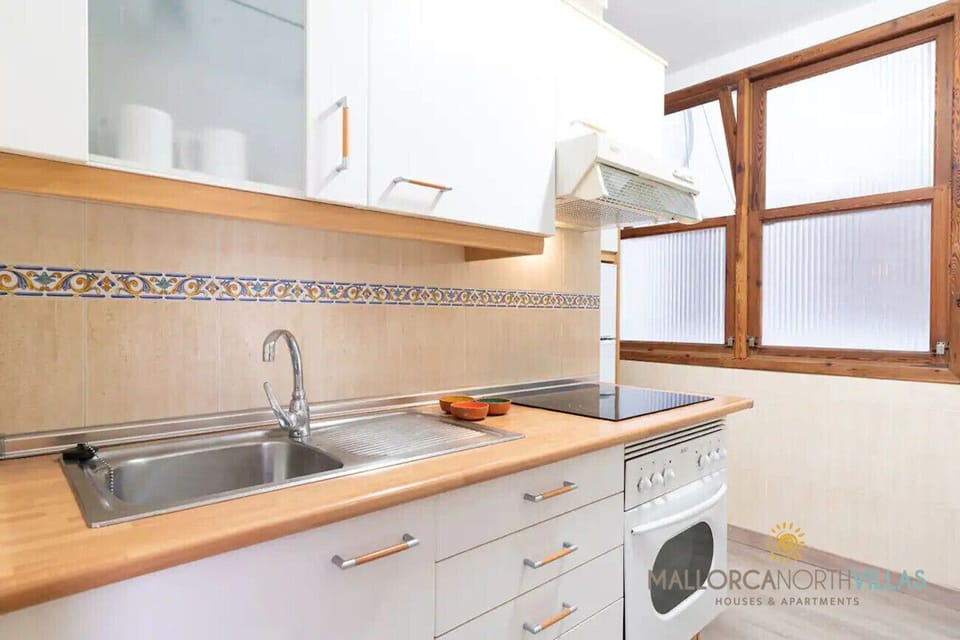Close-up of the kitchen with wooden worktop, stainless-steel sink, ceramic hob, extractor hood, white cabinets and wooden-framed frosted windows with decorative tile border.