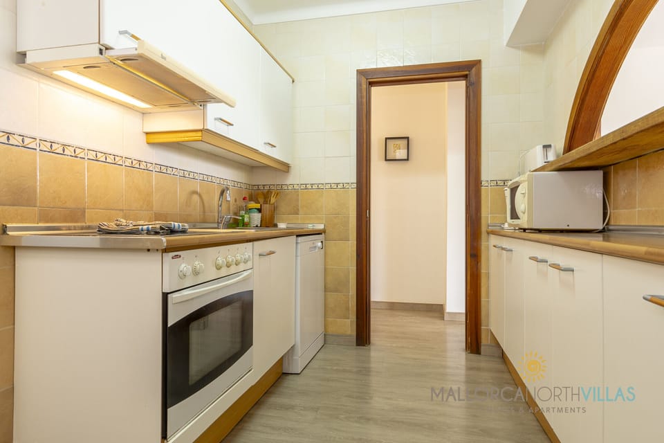 Bright, elongated kitchen with white cabinets, wooden countertop, oven, dishwasher and a spacious work area, connected to the hallway through a wooden door.