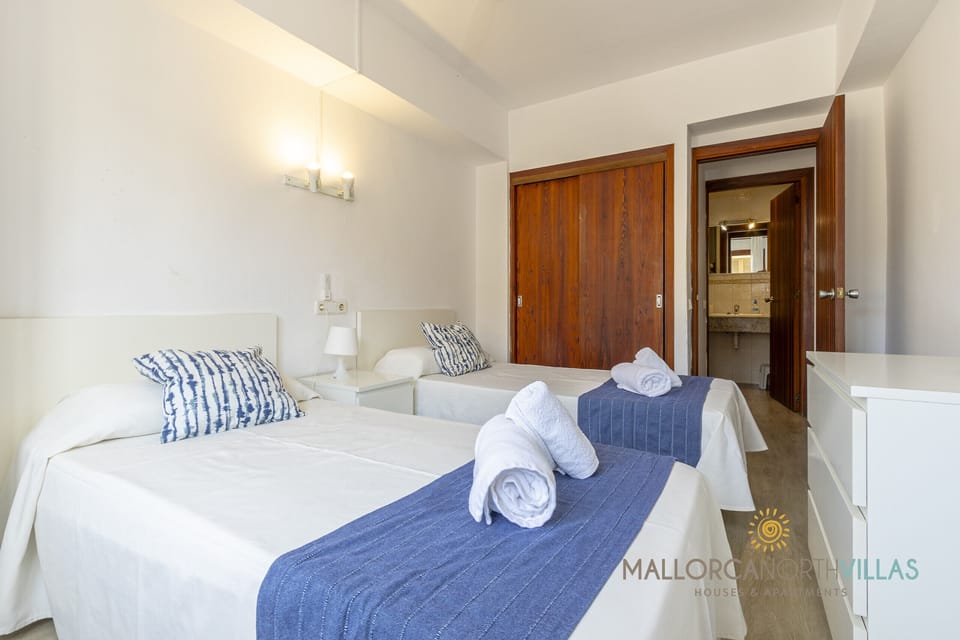 Spacious and bright bedroom with two single beds, white and blue decor, built-in wooden wardrobe and direct access to an en-suite bathroom with a granite countertop.