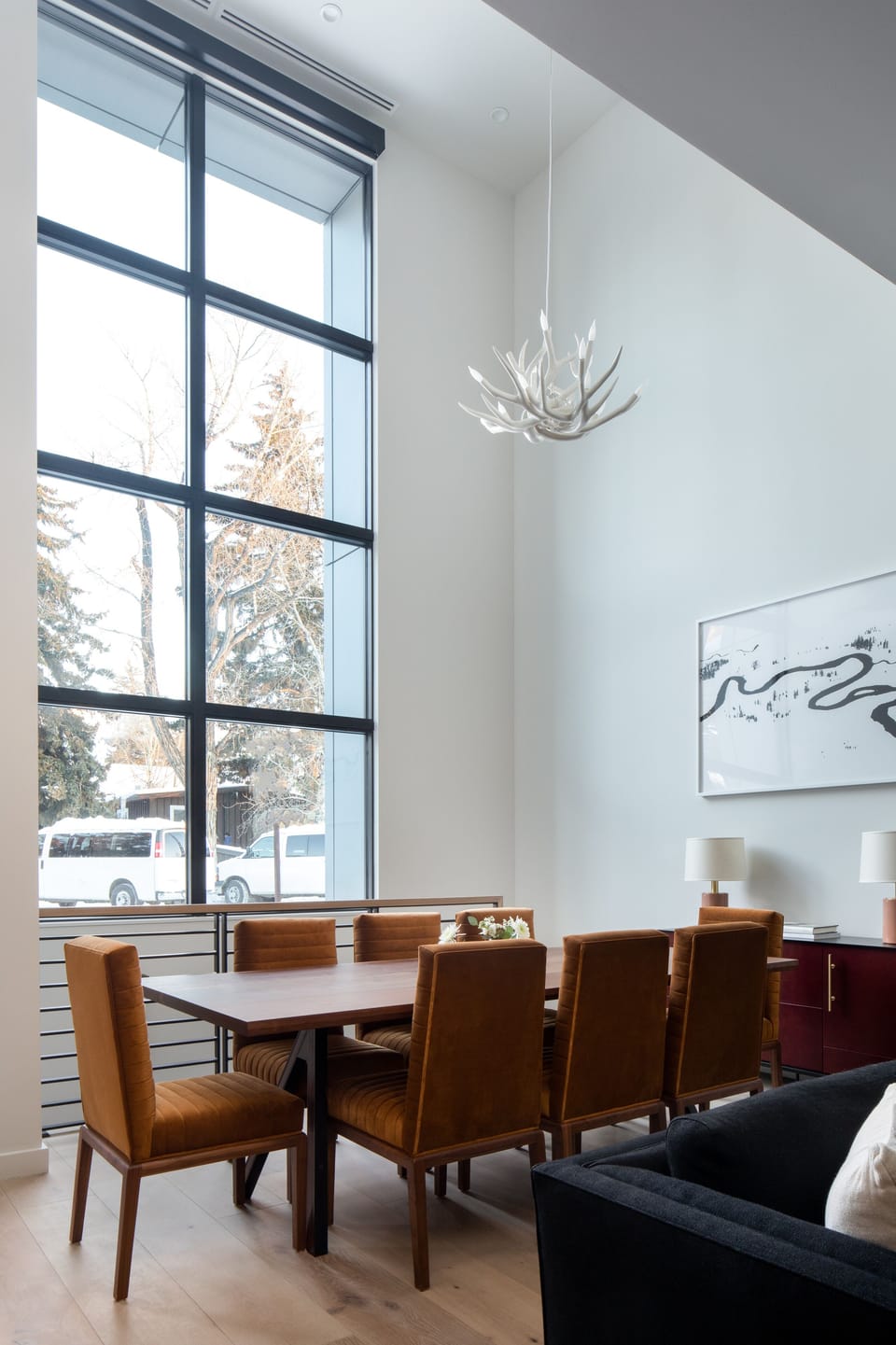 Your elegant dining space awaits with warm leather chairs and soaring windows that frame winter views, where natural light dances through the modern chandelier above your table