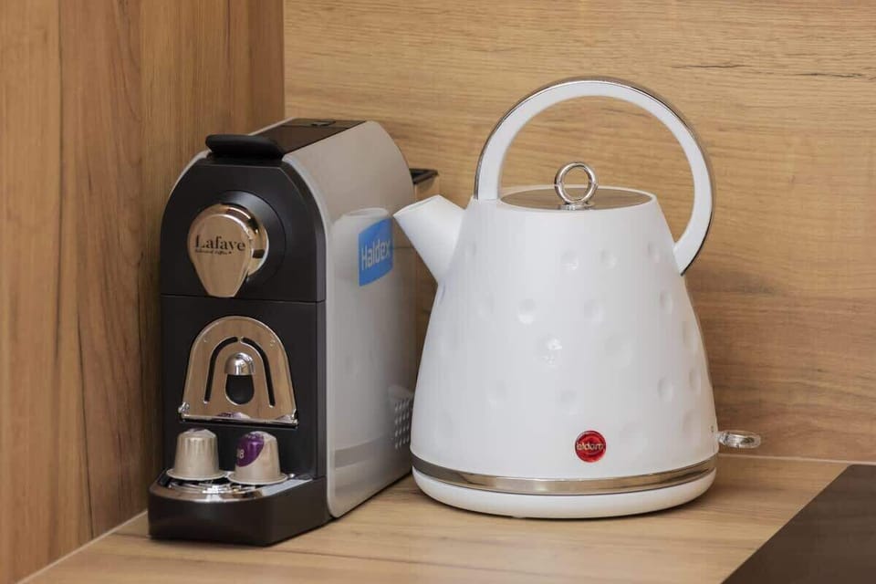 A white electric kettle sits on a wooden kitchen counter. The kitchen has minimalist elements with clean lines and functional design.