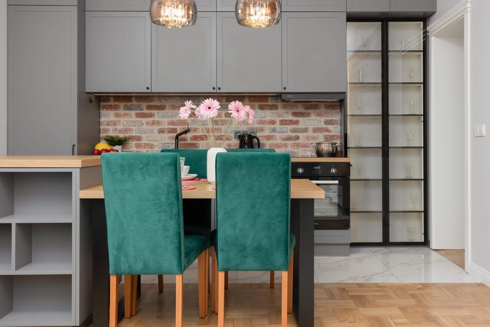 A close-up of a dining table paired with green velvet chairs and pink flowers in a vase.