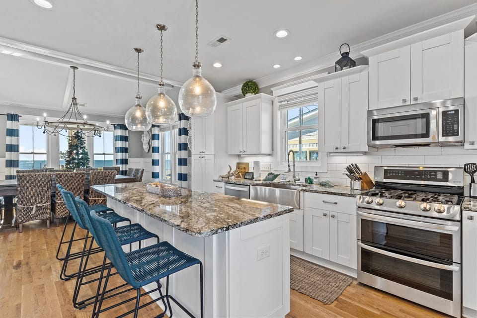 Bright coastal kitchen with white cabinetry, granite island, blue barstools, stainless appliances, pendant lighting, and deck access.