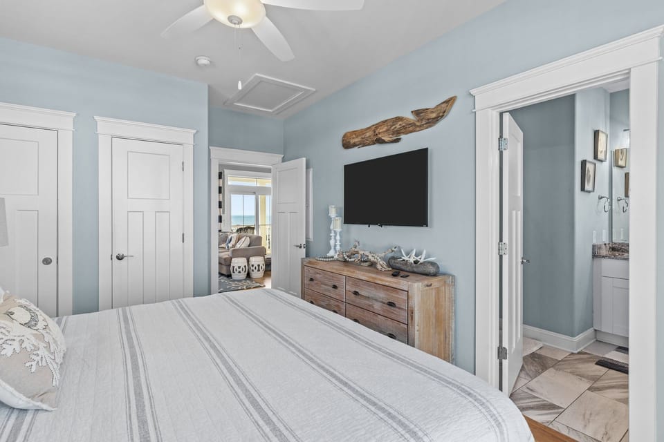 Soft blue bedroom with a TV, wood dresser, driftwood decor, ceiling fan, and easy access to a private ensuite bath.