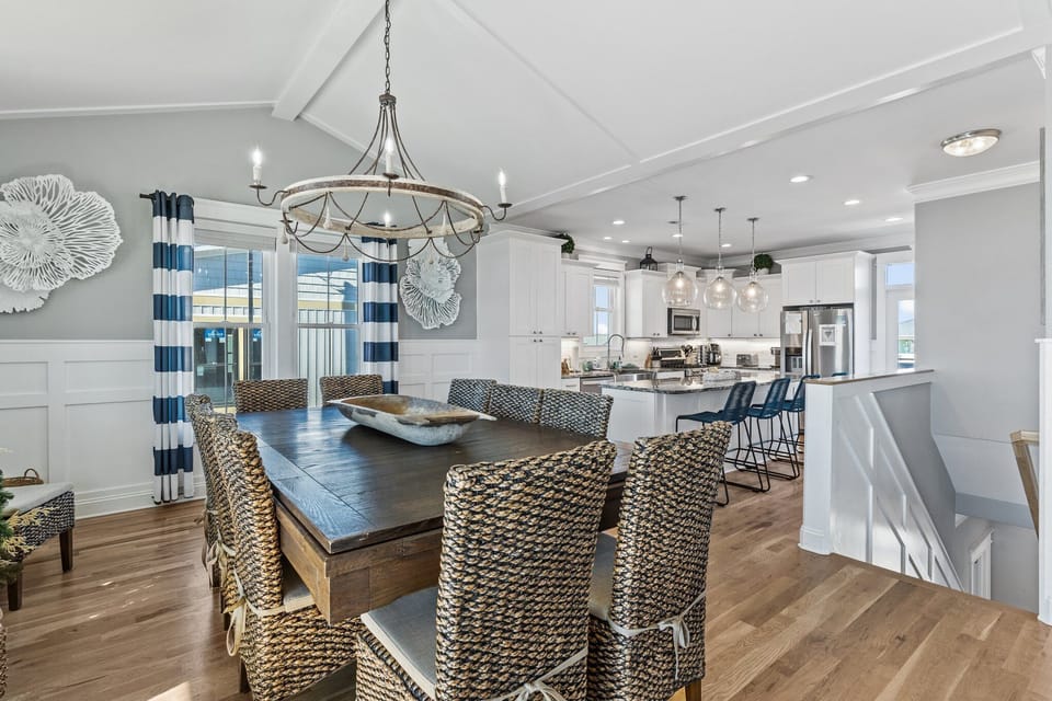 Spacious dining room with seating for eight, woven chairs, hardwood floors, striped curtains, deck access, and bright ocean views through surrounding windows.