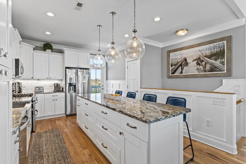Bright coastal kitchen with white cabinetry, granite island, blue barstools, stainless appliances, pendant lighting, and deck access.