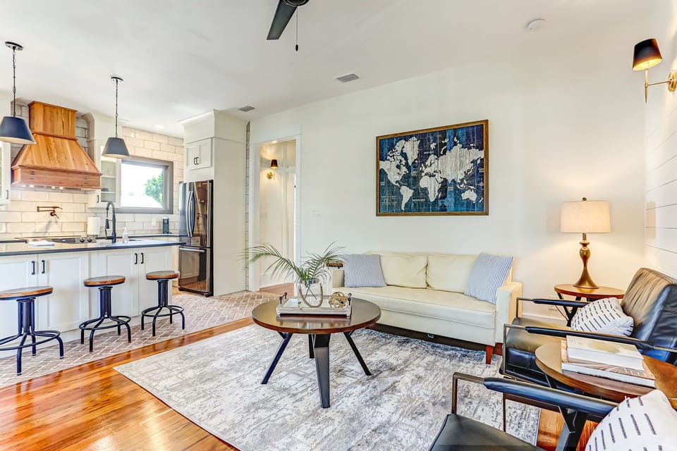 - Enjoy seamless open concept living with this cozy lounge and view of the stylish kitchen
- Relax on the cream sofa or sip coffee at the kitchen island with rustic barstools
- Sunny windows, wood floors, and pendant lights add charm