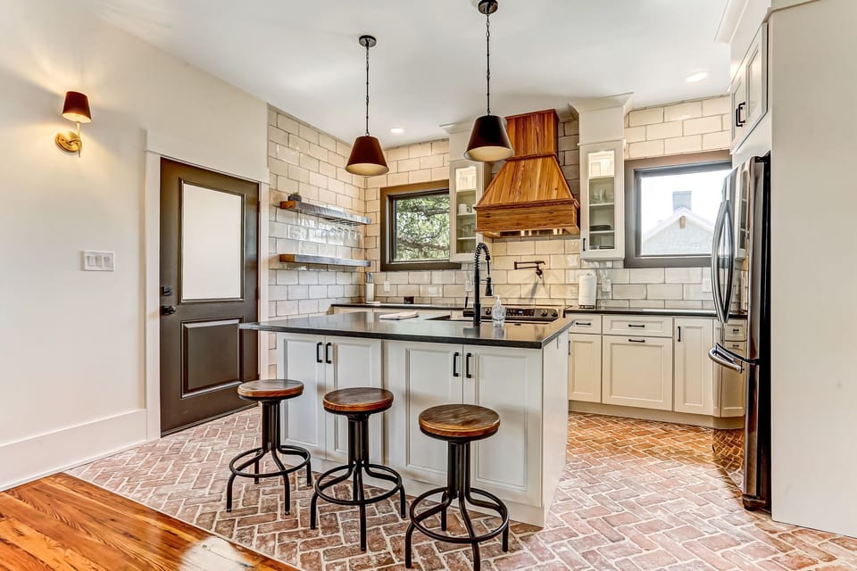 - Cook with ease in this modern kitchen featuring stainless appliances and a farmhouse sink
- Exposed brick floors and wood range hood bring rustic elegance to the space
- Island seating for three under warm pendant lighting invites conversation